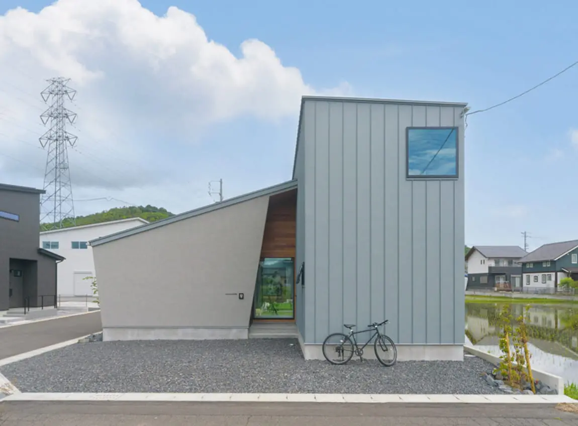 斜めの屋根と直線的なデザインが特徴のモダンな住宅外観。左側はグレーの左官壁、右側は縦張りのガルバリウム外壁で構成され、前には自転車が置かれている。