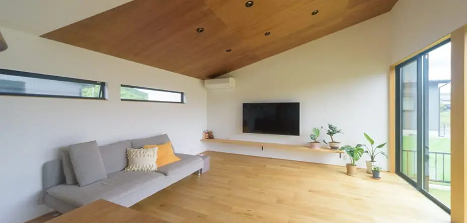 木のぬくもりを感じるリビング。勾配天井に間接照明、フローリングの床にグレーのソファと観葉植物が配置され、壁掛けテレビのある開放的な空間。