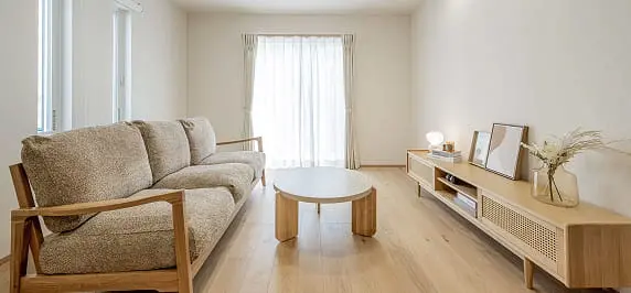 柔らかい色合いのソファと木製家具が配置されたリビングルーム。自然光がレースカーテン越しに差し込み、落ち着いた空間を演出している。