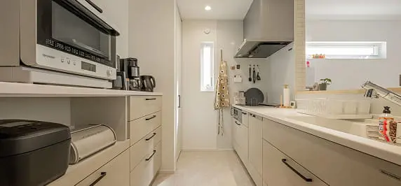 機能的に整えられたキッチン空間。白とグレーの収納、調理家電、吊り戸棚が効率よく配置されており、奥には細長い窓から自然光が差し込んでいる。