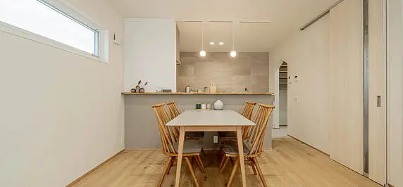 シンプルで温かみのある木製ダイニングセットが置かれたダイニング空間。奥にはグレーのアクセント壁を持つ対面式キッチンが見える。