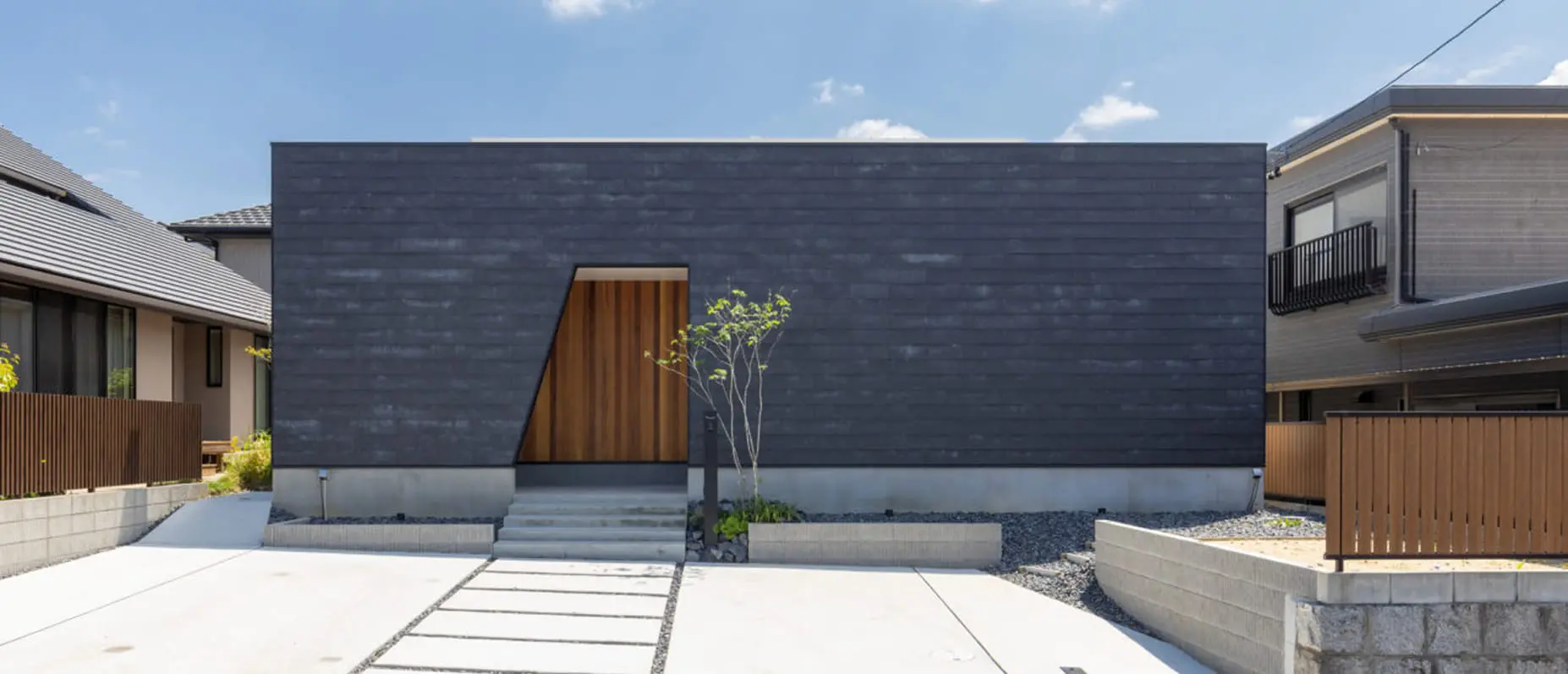 ダークグレーの外壁と木目調の玄関ドアが印象的な平屋住宅。シンプルでミニマルなデザインが特徴で、直線的なファサードがモダンな印象を与える。
