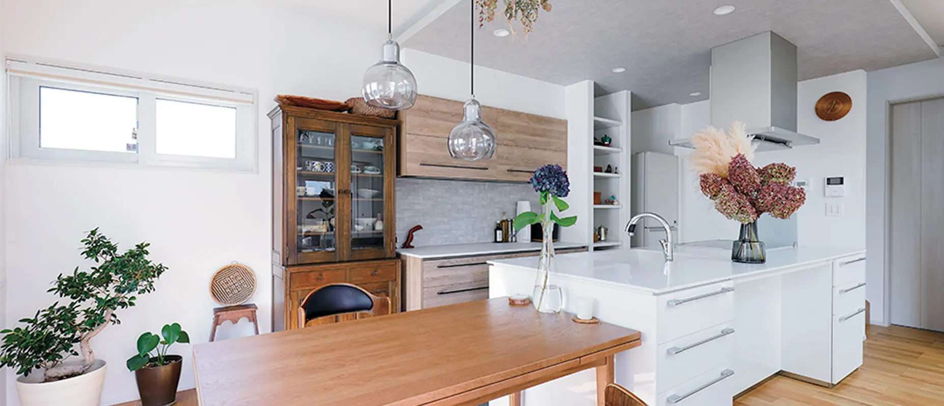 北欧風インテリアで統一された明るいキッチンとダイニング。木製のテーブルとキャビネット、白を基調としたアイランドキッチン、花や観葉植物が彩りを添えている。