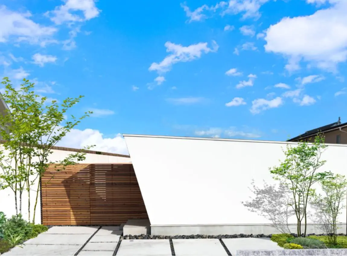 青空の下に建つ、スタイリッシュでモダンな平屋住宅の外観。白い外壁と木目のアクセントが特徴的で、前面には植栽と石を使ったシンプルな外構デザインが施されている。