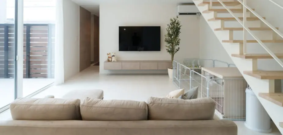 ナチュラルテイストの明るく広々としたリビング。大型ソファの正面に壁掛けテレビがあり、階段下にはペットサークルと観葉植物が配置されている。