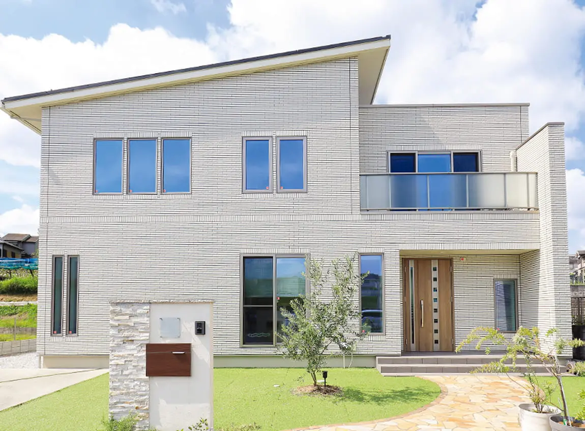 モダンでシンプルなデザインの二階建て住宅の外観。大きな窓とスラント屋根が特徴で、前面には植栽と石畳のアプローチがある。